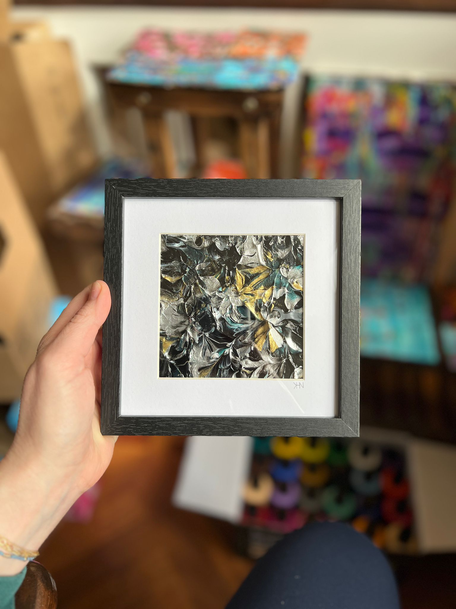 A small framed KNH abstract artwork with black, silver, gold, and teal texture.