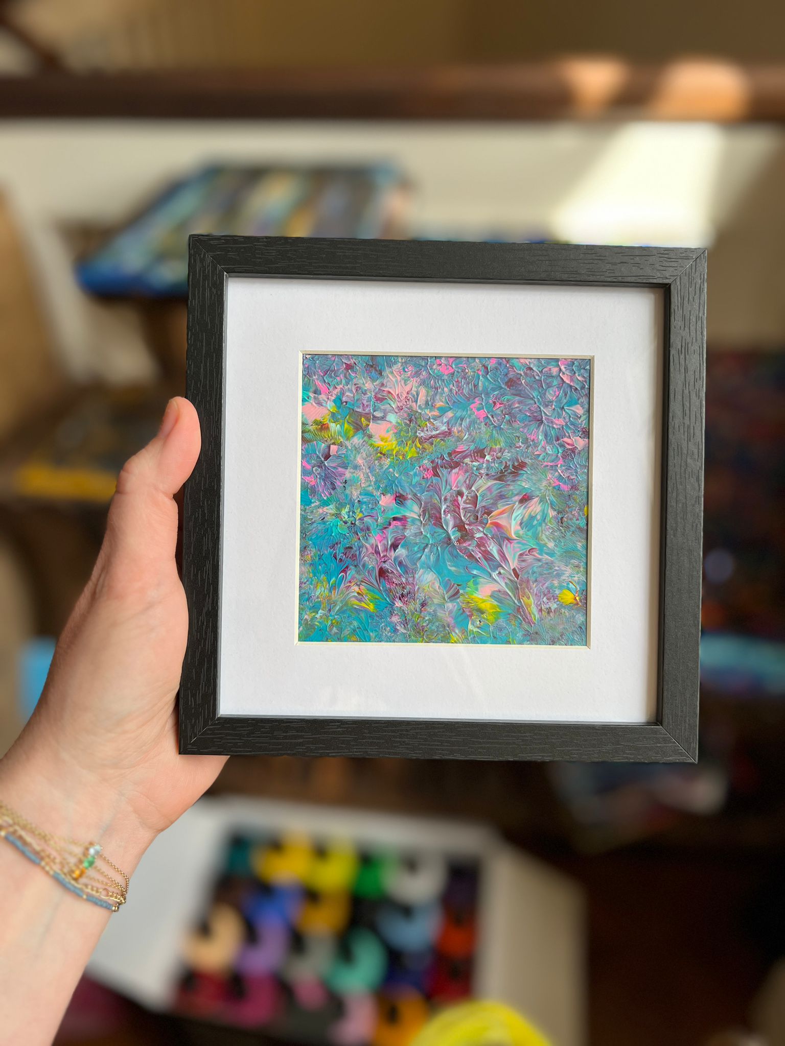 A small framed KNH abstract artwork with turquoise, pink, yellow, and silver texture in a dark frame.